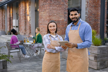 A diverse pair of restaurant staff members smile at the camera outside, while customers dine in the background.