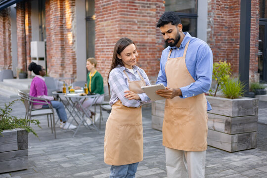 A diverse pair of restaurant staff using a tablet while discussing something outside a brick building.