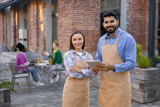 A diverse pair of restaurant staff members smile at the camera outside, while customers dine in the background.