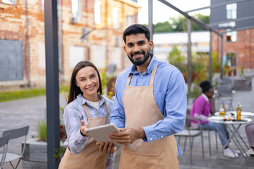 Smiling cafe workers holding a tablet and standing outdoors, with customers in the background.
