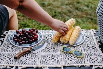 A summer picnic scene featuring corn on the cob, cherries, and vintage green sunglasses. Close-up of a person enjoying a summer lunch or brunch outdoors with corn on the cob, a knife. Summertime fun.