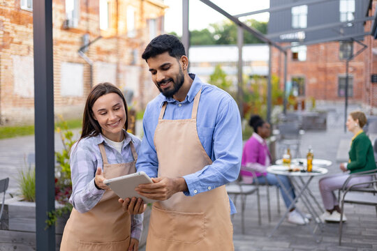 Two restaurant employees reviewing a tablet outdoors, smiling while discussing customer orders in a cafe setting.
