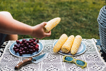 A summer picnic scene featuring corn on the cob, cherries, and vintage green sunglasses. Close-up of a person enjoying a summer lunch or brunch outdoors with corn on the cob, a knife. Summertime fun.