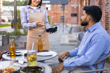 A female waiter offers a payment terminal to a customer at an outdoor restaurant table.