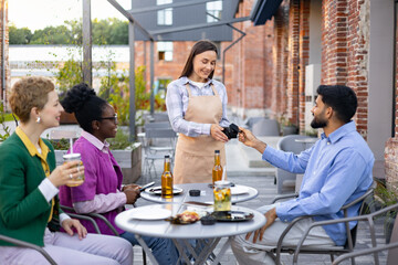 A female waiter assists customers with a portable terminal for payment at an outdoor cafe.