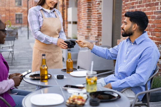 A female waiter at an outdoor cafe assists a customer with payment using a portable terminal.