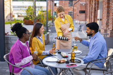 A server presents sushi to a group of friends sitting at an outdoor restaurant table, drinks and food are visible.