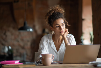 Mujer joven pensativa con cabello rizado, mirando la pantalla de su portátil en casa. Reflexión y concentración en el trabajo remoto, destacando el empoderamiento femenino profesional.