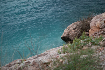 Blue-green ocean water meeting rugged rocky cliffs covered with sparse vegetation, creating a...