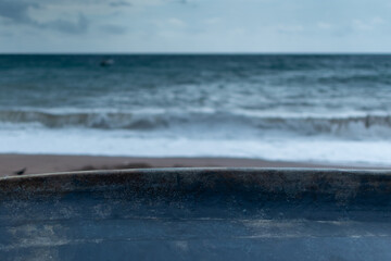 Dark weathered boat rests on sandy beach with ocean waves crashing in the background under cloudy sky