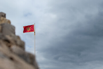 Vibrant red flag of Montenegro adorned with its golden coat of arms, proudly waving on flagpole against backdrop of dramatic cloudy sky and blurred stone structures in the foreground