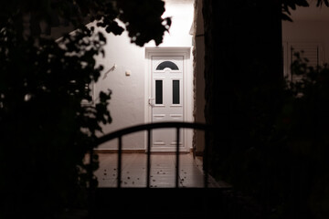 White front door of building brightly lit and framed by shadowy foliage and arching fence creating...