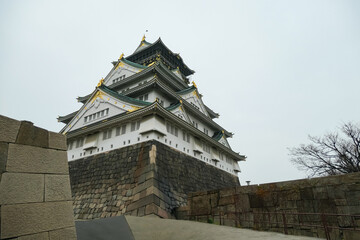 Upward-looking perspective of the majestic Osaka Castle emphasizes its impressive scale and historic stone fortifications, showcasing the grandeur of this iconic Japanese landmark against a grey sky.