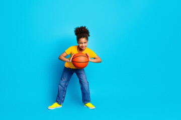 Energetic young girl holding a basketball, dressed in casual clothing in front of a vibrant blue background