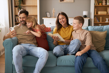 Happy parents enjoying with their kids on sofa in the living room. Mother and son are reading the...