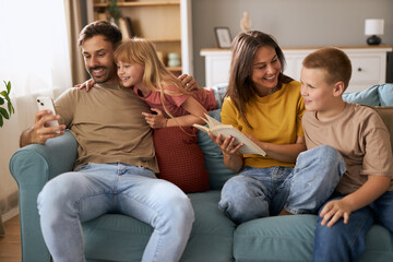 Happy parents enjoying with their kids on sofa in the living room. Mother and son are reading the...