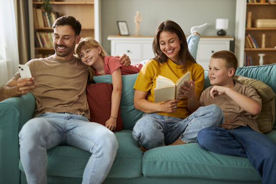 Happy parents enjoying with their kids on sofa in the living room. Mother and son are reading the book, while father and daughter are using cell phone.