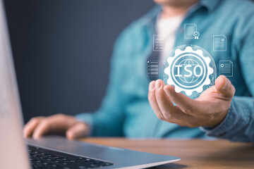 person using a laptop hold a glowing ISO emblem surrounded by digital document icons, symbol certificate standards, quality assurance, regulatory compliance, and international business certifications.