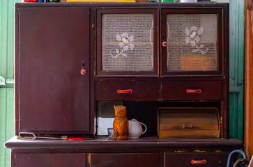 Dark brown kitchen sideboard from the communist era, with lace curtains behind glass doors, a...