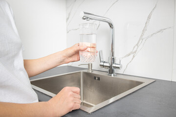 Woman holding a glass pours water from a faucet in the kitchen