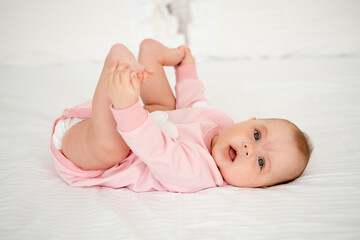 A baby girl plays with her legs lying on her back, a cute and smiling little child in a pink...