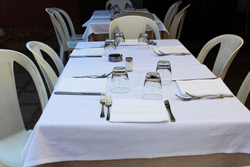 Outdoor dining table set with white tablecloth, cutlery, glassware, simple plastic chairs and a...
