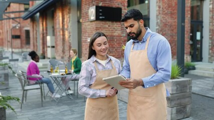 A diverse pair of restaurant staff members smile at the camera outside, while customers dine in the background.
