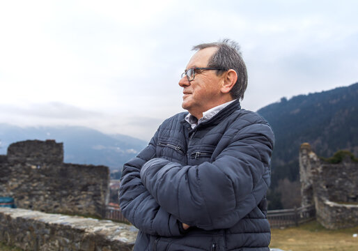 Elderly caucasian male in winter jacket posing near ancient ruins in mountainous landscape - Powered by Adobe