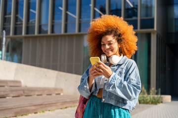 Latin student girl using cellphone phone standing at university campus. Smiling young african american woman with curly ginger hair using smartphone mobile app for studying outdoors. Copy space