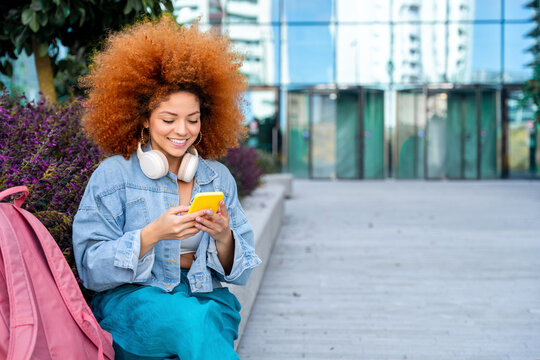 Smiling young african american woman with curly ginger hair using smartphone mobile app for studying outdoors. Latin student girl using cellphone sitting at university campus with backpack. Copy space