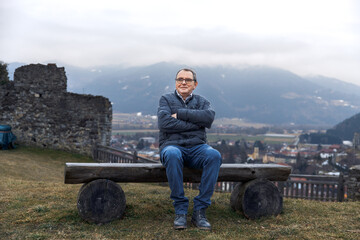 Elderly caucasian man sitting on bench with mountain view and historic ruins