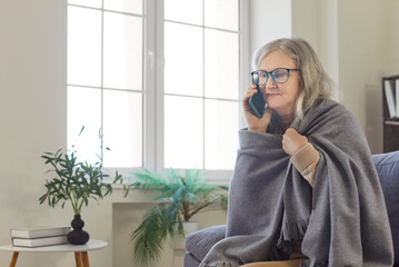 Serious senior woman covered in cozy blanket sitting on home sofa and calling on mobile phone. Old...