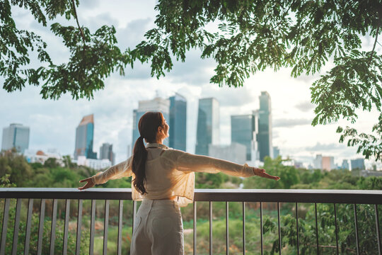 Rear view of happy asian woman wearing headphones enjoying peaceful moment while looking at city skyline with trees and sunset – urban nature, green living and model city lifestyle in public park