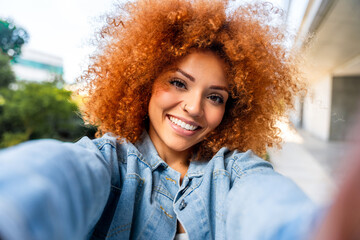 Smiling young african american woman with curly ginger hair using smartphone mobile for selfie, looking at camera outdoors. Student girl using cellphone phone for social media messenger appication