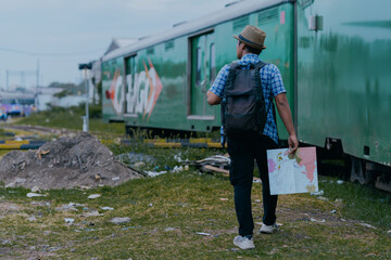 Rear view of asian man in backpack standing near a train with city map in hands and checking his route.