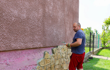 A man meticulously lays decorative stone on the house foundation. He uses a trowel to apply mortar,...