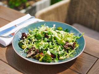 Fresh green salad in plate made of green leaves mix, broccoli and tomatoes on wooden table outdoor restaurant. Variety of healthy fresh harvested eatable greens. Raw food, organic, detox diet concept