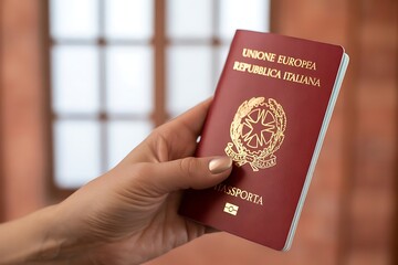 Close up of a hand holding a red italian european union passport with gold lettering and emblem against a blurred background of a window and brick wall