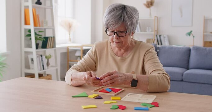 Busy old woman collecting colourful figures, solving puzzle, sitting at deak in living room. Elderly lady enjoying free time, playing educational game. Old people and memory training concept