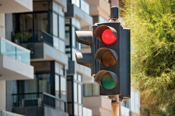Close-up of a dirty traffic light with the red light on and sun visors, set against a modern building with glass balconies and green foliage.