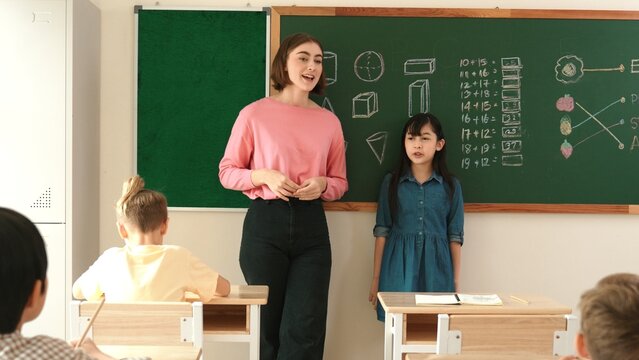 Caucasian teacher asking for volunteer while asian girl walking at in front of class with confident. Smart student raised hand and answer question while teacher clapping hands to encourage. Pedagogy.