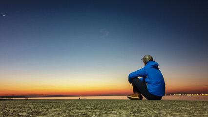Lone man in a blue jacket sits on a pier, contemplating serene scene as the last light of sunset...
