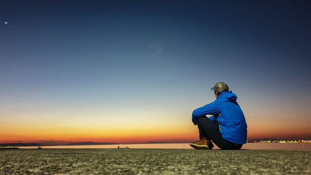 Lone man in a blue jacket sits on a pier, contemplating serene scene as the last light of sunset fades into a deep blue night sky over the water, capturing a moment of quiet introspection and peace.