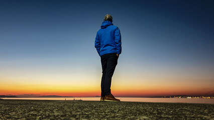 From low angle, man in a blue jacket stands alone on pier, looking out at distant city lights...