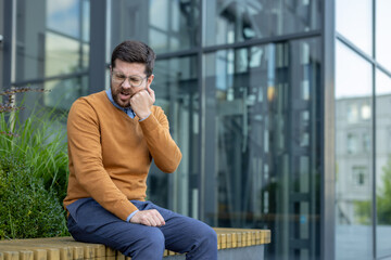 A young male businessman sits on a bench near an office building and holds his cheek with his hand,...
