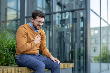 A young man, twisted in pain, sits on a bench near an office building and holds his hand to his...