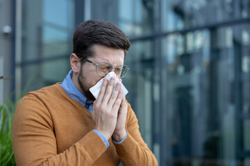 A close-up photo of a young man sitting near an office center on a sidewalk wiping his runny nose...