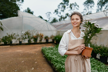Young female gardener in apron smiling while holding a potted plant in a greenhouse, showcasing...