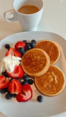 Fluffy pancakes served with strawberries, blueberries, cream, and a cup of coffee on a white plate. This image represents a cozy morning breakfast and sweet indulgence concept