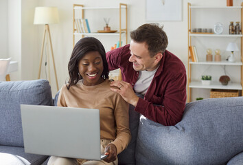 Fototapeta premium Smiling and cheerful married couple using laptop computer for online entertainment. Talking and relaxing at home living room. Internet leisure and happy moments in a cozy domestic setting.